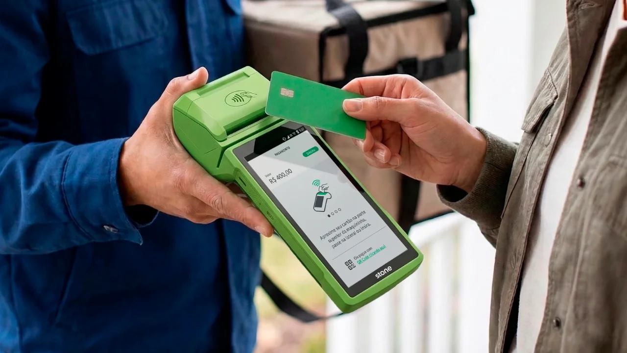 Close-up de um entregador segurando uma maquininha de cartões verde da Stone, enquanto um cliente aproxima um cartão de crédito verde para realizar um pagamento por aproximação. Ao fundo, uma mochila de entregas desfocada.
