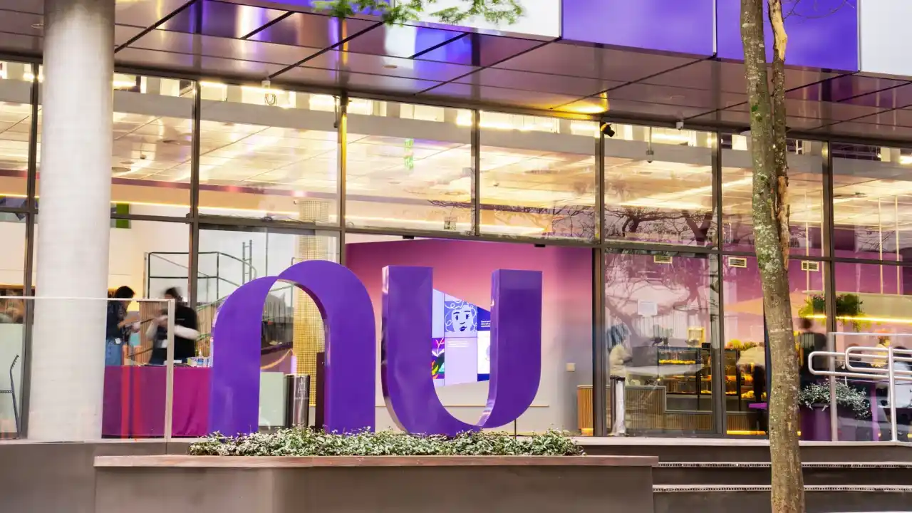 Fachada moderna de uma unidade do Nubank em um entardecer. Em primeiro plano, o logotipo "nu" gigante e roxo posicionado sobre um canteiro de plantas. Ao fundo, através das grandes janelas de vidro iluminadas, nota-se o interior do escritório com pessoas circulando e um balcão de cafeteria. A arquitetura é contemporânea, com painéis metálicos roxos e brancos no teto externo.