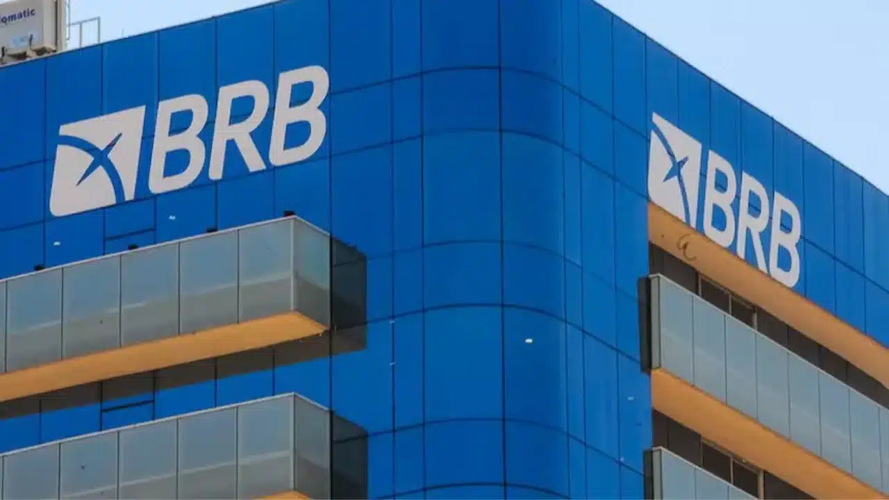 Fotografia em ângulo baixo da fachada moderna do edifício sede do Banco de Brasília (BRB). O prédio possui revestimento em vidro espelhado azul e exibe o logotipo branco e azul do BRB em destaque no topo da estrutura. O céu está limpo ao fundo.