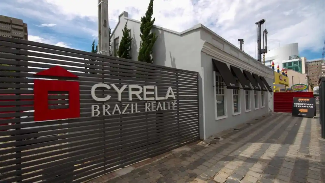 Fachada de um edifício corporativo moderno e branco da MBRF Global Foods Company. O logotipo azul da MBRF está em destaque na parede lateral direita. A entrada possui grandes painéis de vidro escuro sob uma cobertura cinza metálica. O céu está azul limpo com poucas nuvens, e há um jardim baixo e palmeiras decorativas na frente.