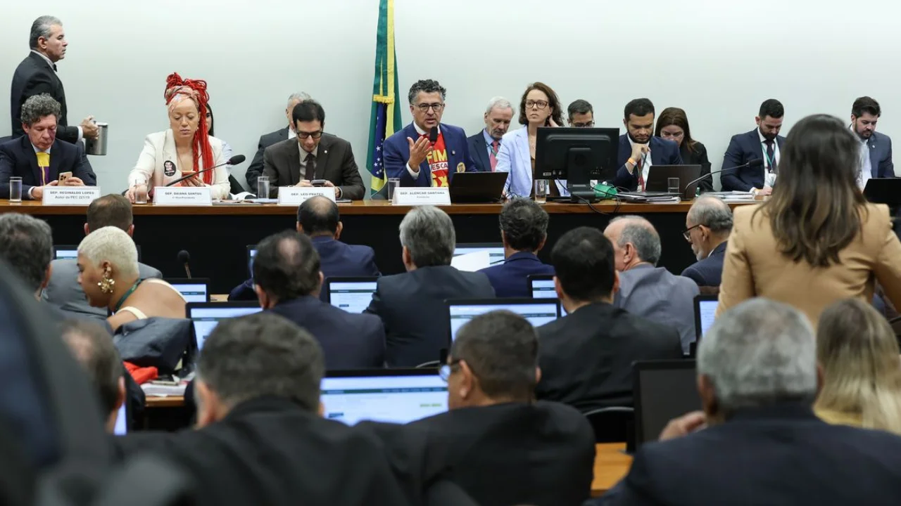 Vista de uma mesa de presidência em comissão da Câmara dos Deputados. No centro, o deputado Alencar Santana fala ao microfone vestindo uma camiseta vermelha com os dizeres "FIM DA ESCALA 6X1". À sua esquerda, a deputada Daiana Santos e outros parlamentares analisam documentos e notebooks. A plateia, composta por assessores e deputados de costas, observa a sessão. Bandeiras do Brasil são visíveis ao fundo da sala.
