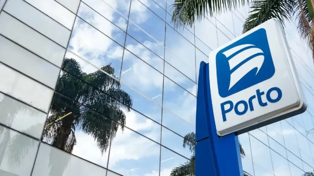 Totem azul e branco com o novo logotipo da Porto em destaque à direita. Ao fundo, a fachada de vidro de um edifício corporativo moderno reflete palmeiras e um céu azul com nuvens claras.