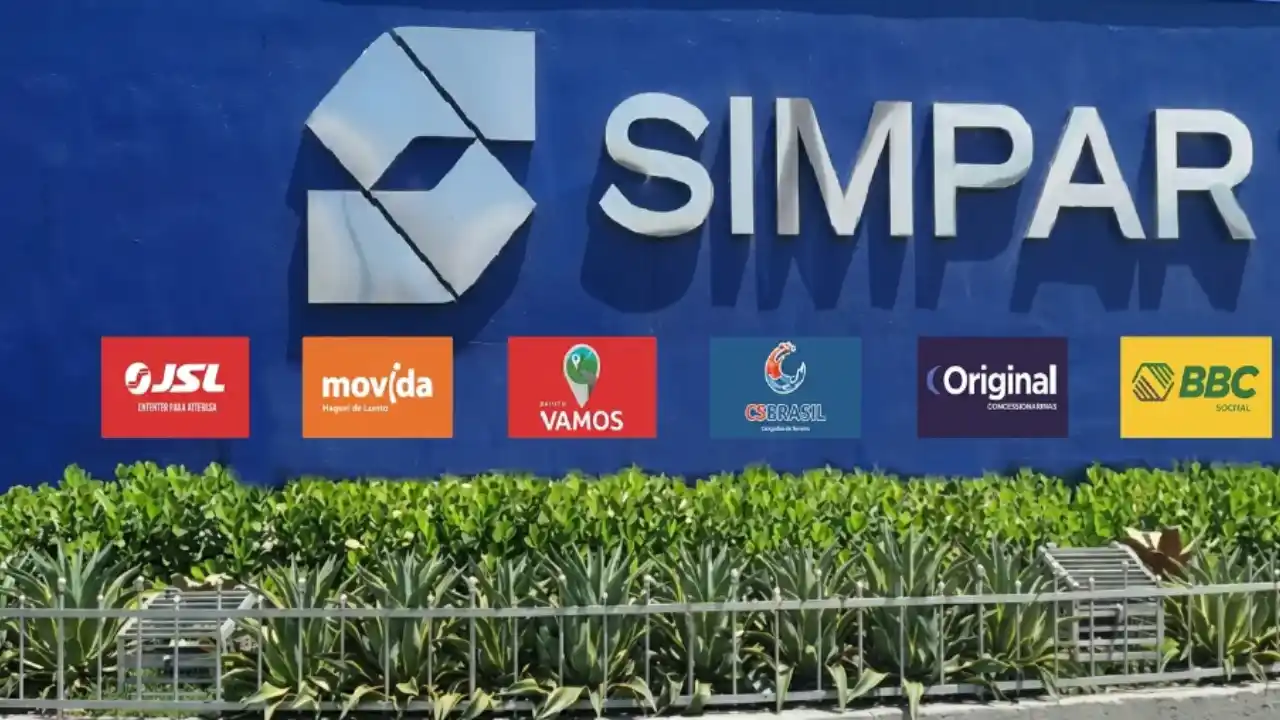 achada azul com o logotipo prateado da Simpar em destaque na parte superior. Abaixo, uma sequência de placas coloridas exibe os logotipos das empresas do grupo: JSL (vermelho), Movida (laranja), Vamos (vermelho e verde), CS Brasil (azul), Original (roxo) e BBC (amarelo). À frente, há um jardim com plantas verdes e uma grade metálica.