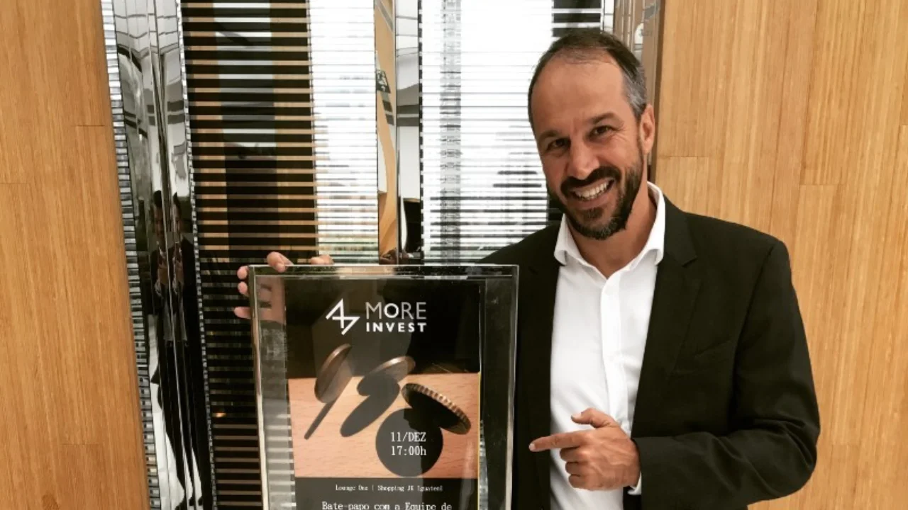Fotografia de plano médio de Carlo Moratelli, um homem de pele clara, cabelos curtos escuros e barba aparada, vestindo camisa social branca aberta no colarinho e paletó preto. Ele sorri para a câmera enquanto segura com a mão direita um cartaz emoldurado com o logotipo "More Invest" e informações sobre um bate-papo no Shopping JK Iguatemi. Com o dedo indicador esquerdo, ele aponta para o anúncio. O fundo exibe uma parede com textura de ripas verticais de madeira e detalhes espelhados.