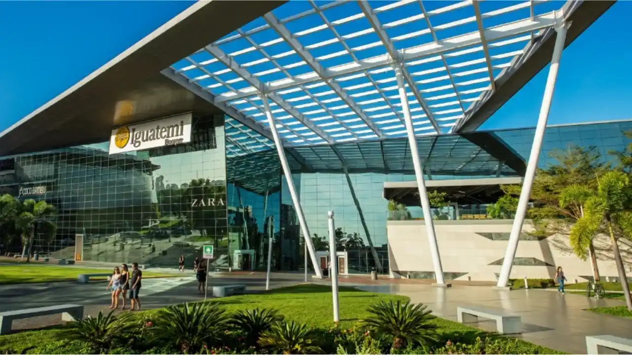 Fotografia em perspectiva angular da entrada principal do shopping center Iguatemi Bosque. A arquitetura moderna destaca-se por uma gigantesca estrutura de cobertura em treliça metálica branca, disposta de forma geométrica e sustentada por grossas colunas inclinadas sobre um pátio externo. A fachada principal do edifício é inteiramente revestida por painéis de vidro espelhado azulado, refletindo o céu limpo. No topo esquerdo da fachada, encontra-se o letreiro iluminado "Iguatemi Bosque" com seu logotipo circular amarelo. Através das vidraças, nota-se a identificação interna da loja Zara. No plano inferior, um gramado verde com arbustos e palmeiras ornamentais margeia a calçada onde alguns pedestres caminham sob a luz forte do dia.