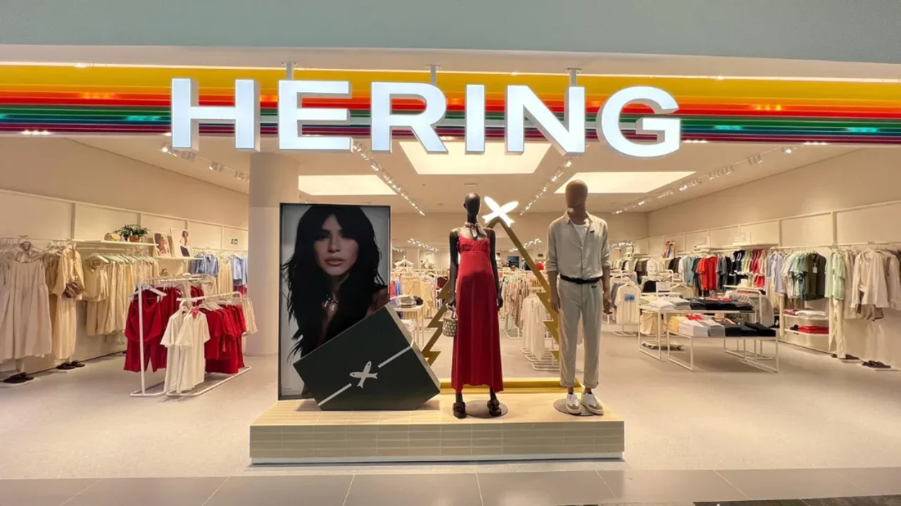 Fotografia em plano frontal da fachada iluminada de uma loja de roupas Hering em um shopping center. No topo, o logotipo com a palavra "HERING" em letras brancas e maiúsculas destaca-se sob uma testeira decorada com faixas horizontais coloridas em tons de amarelo, laranja, vermelho, rosa, azul e verde. Na vitrine central e plataforma de exposição aberta, há dois manequins estilizados: uma silhueta feminina preta vestindo um longo vestido vermelho de alças e um manequim masculino bege com traje casual claro. Ao lado, um totem exibe a foto em close de uma modelo de cabelos escuros e, no chão, uma grande caixa decorativa preta traz o símbolo estilizado de dois peixes cruzados da marca. O interior da loja aparece amplo, claro e organizado ao fundo.