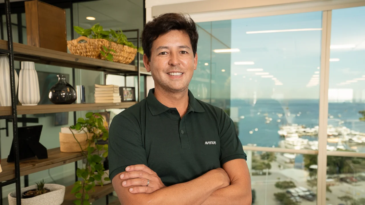 Fotografia em plano médio de Roberto Lee, CEO da Avenue. Um homem de cabelos castanhos curtos, sorrindo de braços cruzados no centro da imagem, vestindo uma camisa polo verde-escura com o logotipo "Avenue" bordado em branco no peito. À esquerda, observa-se uma estante de madeira decorada com plantas de folhas verdes em vasos e livros. À direita, ao fundo, uma grande janela de vidro revela uma vista panorâmica ensolarada de uma marina repleta de barcos e o mar azul sob um céu claro.