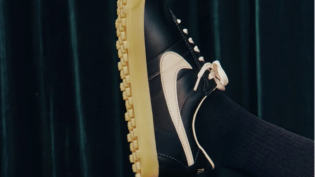 Fotografia em plano médio da fachada moderna e minimalista de uma loja conceito de varejo global da Nike à noite. Em destaque na parede de concreto cinza texturizado, o logotipo "Swoosh" da Nike brilha intensamente em luz branca LED. Amplas vitrines de vidro transparente revelam um interior sofisticado e bem iluminado, com fileiras organizadas de tênis de corrida de alta performance em prateleiras flutuantes. Na calçada em frente à loja, pedestres urbanos desfocados por efeito de movimento (motion blur) caminham sob a iluminação suave dos postes públicos, refletindo o dinamismo comercial da marca.