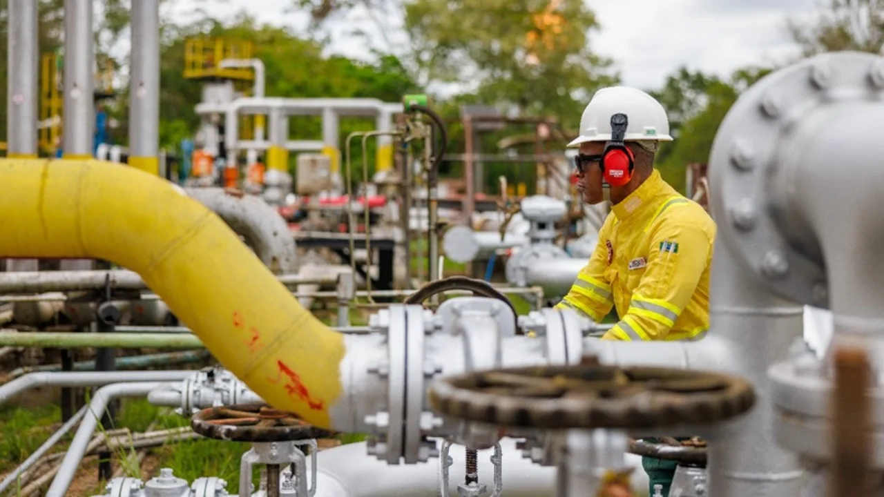 Fotografia em plano médio e ângulo lateral de um operador técnico trabalhando em um campo de petróleo terrestre durante o dia. O profissional veste uniforme amarelo com faixas refletivas, capacete branco e protetores auriculares vermelhos, manuseando conexões industriais. Em primeiro plano, destaca-se uma tubulação amarela curvada. O cenário é preenchido por engrenagens, registros, tubos de metal cinza e vegetação verde ao fundo sob céu nublado.