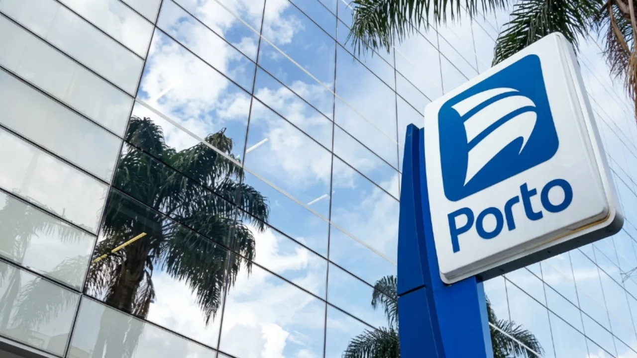 Fotografia em ângulo de baixo para cima (contra-plongé) com foco em uma placa de sinalização vertical azul e branca da Porto Seguro, exibindo o logotipo de três faixas brancas curvadas dentro de um quadrado azul e a inscrição "Porto" logo abaixo. Ao fundo, a fachada de um moderno edifício de escritórios corporativos com janelas de vidro espelhadas reflete com clareza palmeiras e um céu azul com nuvens brancas esparsas.