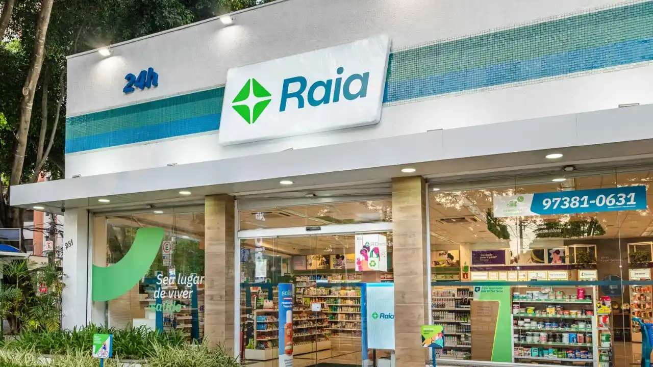 Fotografia em plano aberto e diagonal da fachada moderna de uma farmácia Droga Raia, do grupo RD Saúde. Na parte superior da parede externa branca, destaca-se um grande painel luminoso com o logotipo da marca — uma cruz estilizada de quatro pontas em tons de verde — ao lado do nome "Raia" em letras azuis. À esquerda, vê-se a inscrição "24h" em letras azuis iluminadas. A fachada frontal é composta por grandes vitrines de vidro transparente que revelam o interior da loja muito bem iluminado, com gôndolas e prateleiras organizadas e repletas de produtos. No vidro esquerdo, há um grande adesivo verde com a frase "Seu lugar de viver saúde".