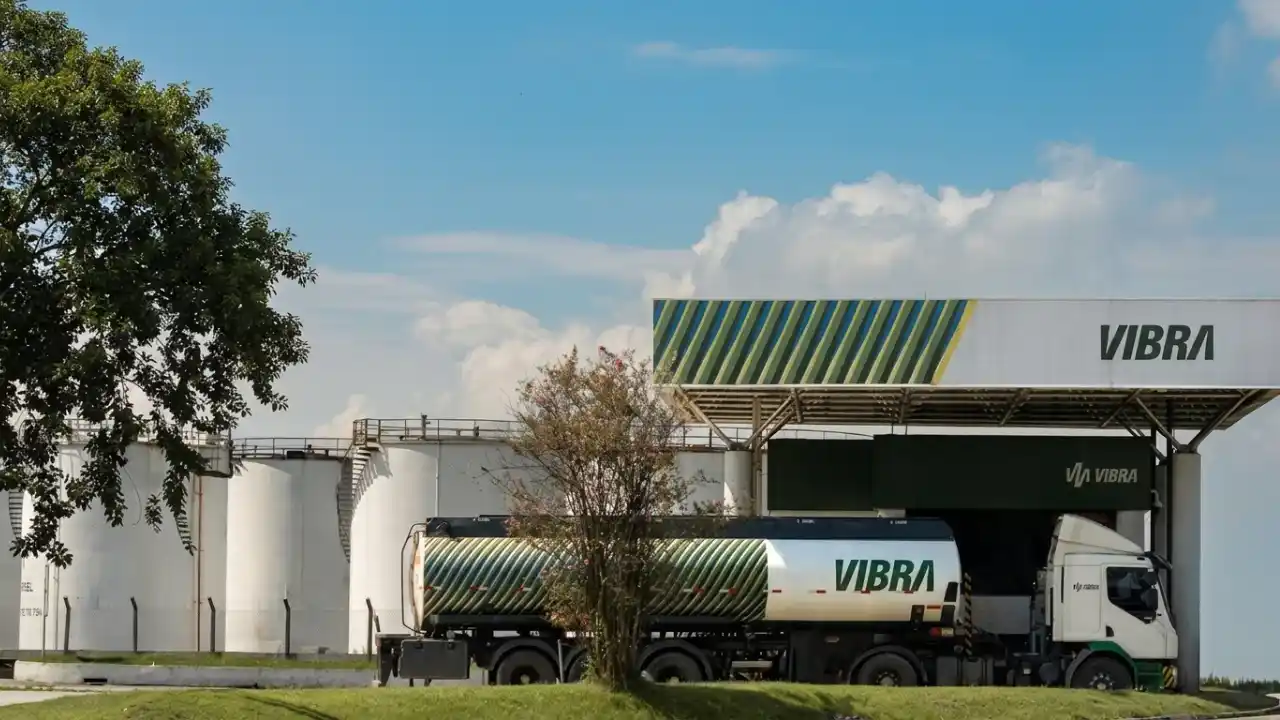 Fotografia em plano aberto de uma base operacional de distribuição de combustíveis da Vibra sob um céu azul com nuvens brancas. À esquerda, veem-se três grandes tanques cilíndricos de armazenamento brancos. Ao centro, um caminhão-tanque branco com listras diagonais verdes e amarelas e o logotipo "VIBRA" em letras verdes está estacionado sob a cobertura de abastecimento de um galpão industrial, que também exibe a marca "VIBRA". Uma árvore de folhas finas aparece em primeiro plano, cobrindo parcialmente a lateral do caminhão.