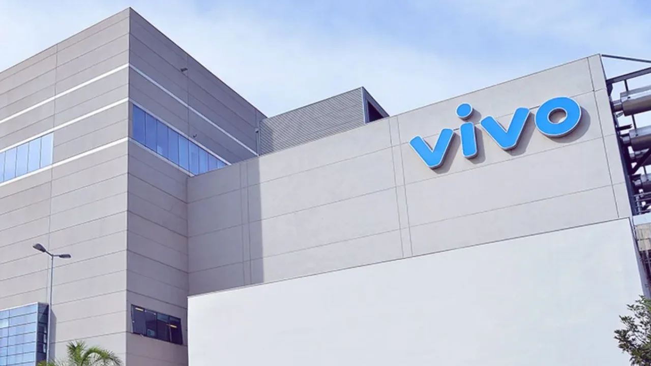 Fotografia em plano médio de ângulo contrapicado mostrando a fachada moderna de um grande edifício corporativo de concreto cinza e branco. No canto superior direito da parede principal, destaca-se o logotipo da marca "vivo" em letras tridimensionais azuis. À esquerda, observam-se janelas de vidro refletivo azul e, na calçada, um poste de iluminação pública com folhas de uma palmeira surgindo na parte inferior. O céu ao fundo apresenta-se claro e limpo.
