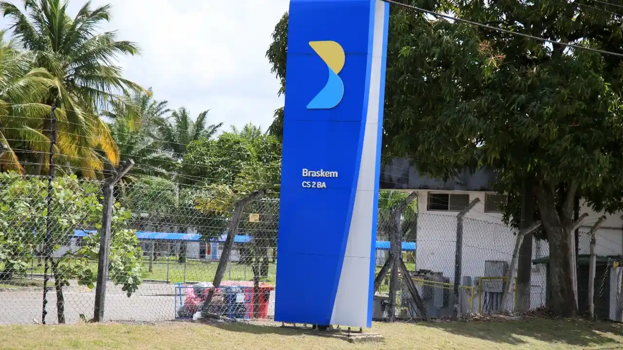 Totem vertical de sinalização em tom azul-escuro exibindo no topo o símbolo estilizado da Braskem em amarelo e ciano, acompanhado das inscrições "Braskem" e "CS 2 BA". A estrutura está posicionada em um gramado ao lado de palmeiras e árvores tropicais, delimitada por uma cerca de tela metálica com prédios industriais e operacionais ao fundo.