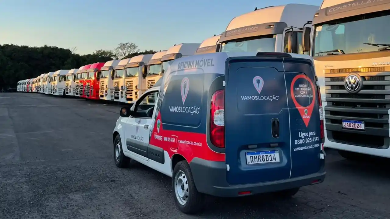 Fotografia em plano médio de um pátio de veículos industriais sob iluminação suave de fim de tarde. Em primeiro plano e de perfil traseiro, uma van utilitária nas cores branca, azul-escuro e vermelha exibe as marcas "VAMOS LOCAÇÃO" com ícones vermelhos de pin de localização e a inscrição "OFICINA MÓVEL" no topo das laterais. Na traseira da van, visualiza-se a placa de identificação "RMR8D14" e informações de contato comercial. Ao fundo, estende-se uma longa e simétrica fileira de caminhões pesados de cabine branca, destacando-se à direita um caminhão Volkswagen Constellation com grade frontal preta, logotipo da marca em destaque e placa "JAU2D24". O pátio é pavimentado em asfalto escuro e há árvores verdes sob o céu limpo ao fundo.