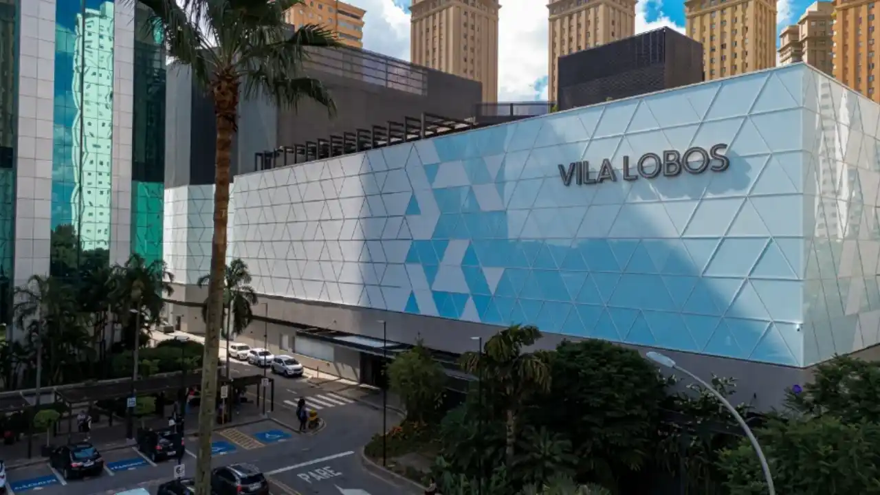 Fotografia em plano médio da fachada moderna do Shopping Vila Lobos em São Paulo, empreendimento gerido pela Allos. A estrutura exibe painel arquitetônico geométrico nas cores azul e branca com o letreiro "VILA LOBOS" fixado na parte superior. À esquerda, ergue-se uma torre corporativa espelhada em vidros esverdeados com palmeiras e coqueiros na calçada. Em primeiro plano, observa-se a via de acesso de veículos e vagas de estacionamento sob céu azul com nuvens suaves.
