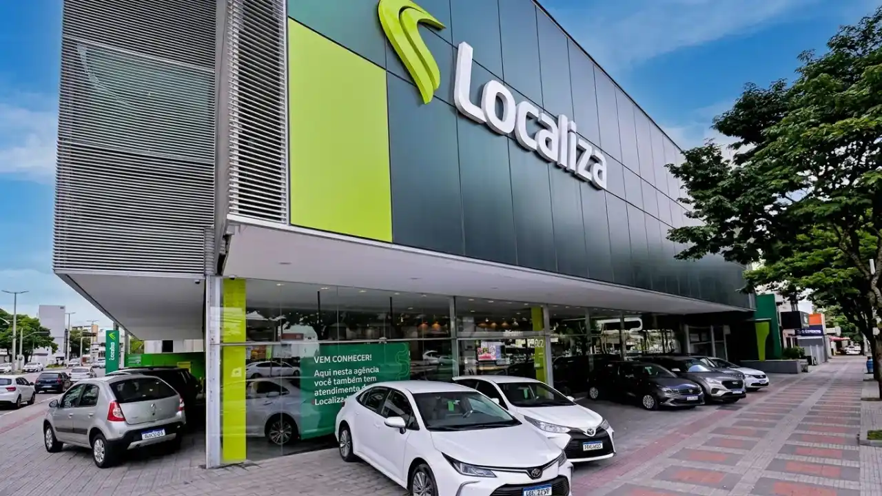 Fotografia em plano aberto e ângulo diagonal da fachada externa de uma grande e moderna agência da Localiza. O edifício apresenta painéis arquitetônicos em cinza-escuro e verde-limão, destacando o logotipo oficial em relevo branco "Localiza" no topo. A base do prédio é fechada por grandes vidraças transparentes, onde há um banner promocional verde com o texto "VEM CONHECER! Aqui nesta agência você também tem atendimento...". Na área de estacionamento frontal, estão alinhados diversos carros seminovos e de locação, incluindo um sedã branco Toyota Corolla em destaque central, um veículo Toyota compacto e um hatch prata na lateral esquerda. Uma calçada com árvores e uma rua aparecem à direita sob um céu claro.