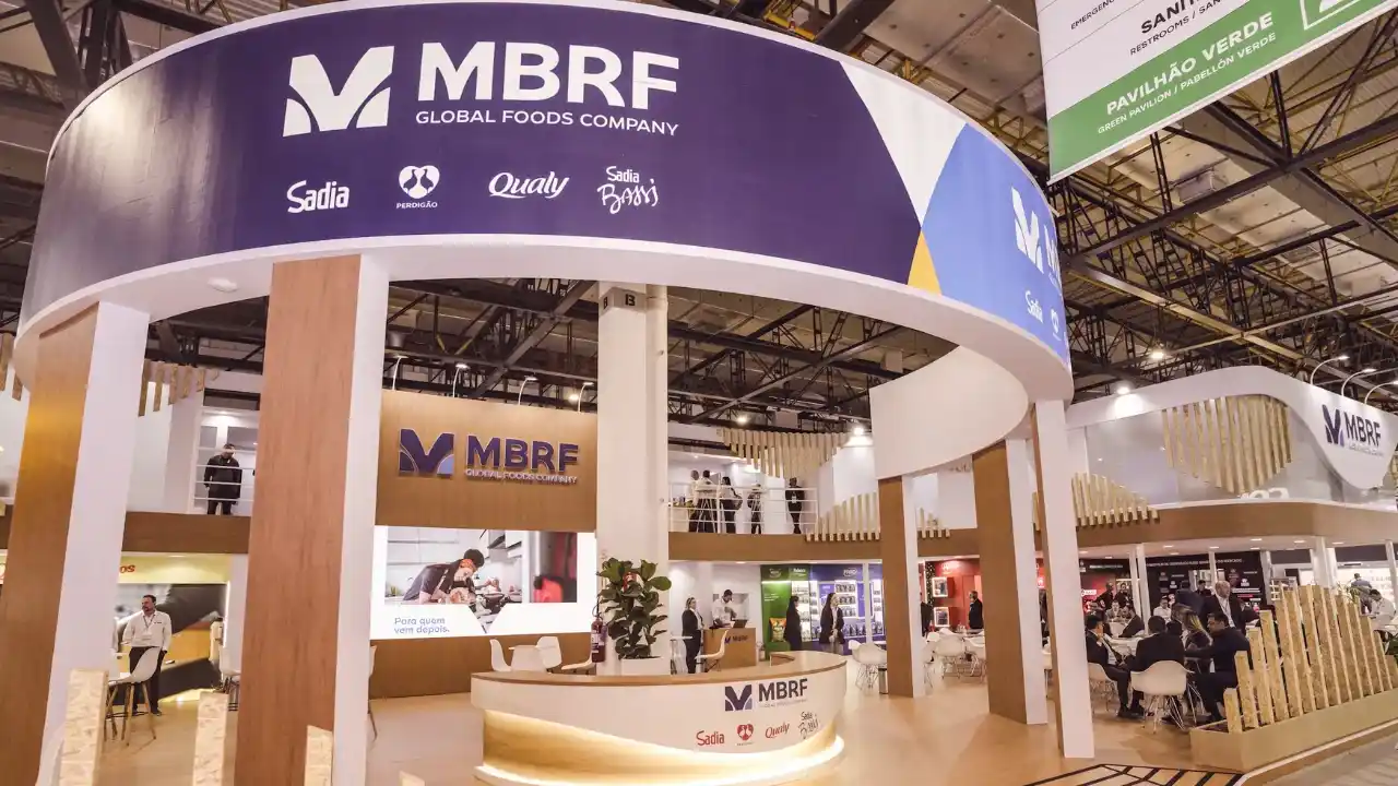 Fotografia em grande-angular do estande de exposições da MBRF Global Foods Company em uma feira do setor de alimentos. O pavilhão apresenta uma estrutura circular suspensa em azul e madeira com a marca "MBRF" em destaque, acompanhada dos logotipos das subsidiárias Sadia, Perdigão, Qualy e Sadia Bio. Abaixo, vê-se um balcão de recepção iluminado, salas de reuniões integradas com móveis modernos e visitantes conversando.