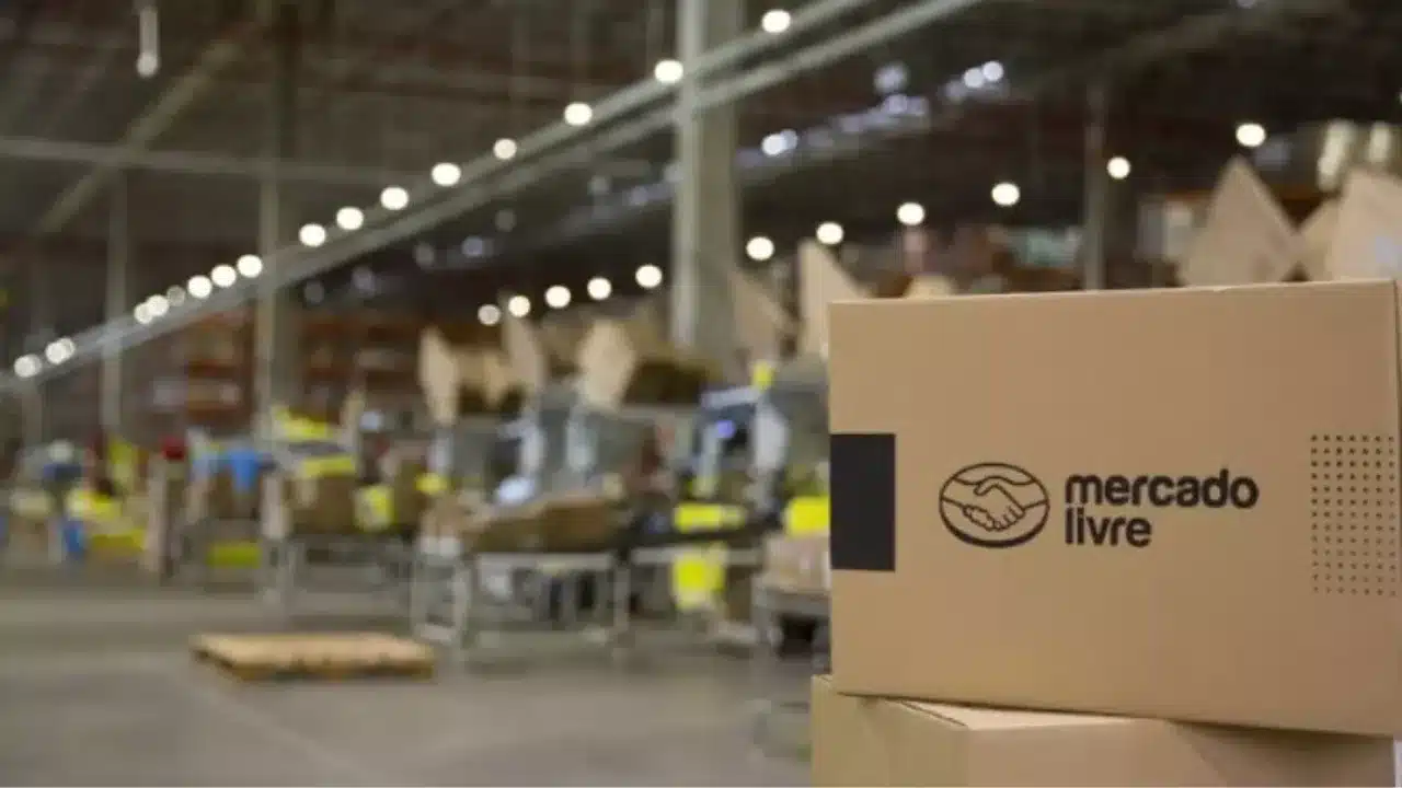 Fotografia em close-up de caixas de papelão empilhadas em primeiro plano, exibindo o logotipo de aperto de mãos e o nome "mercado livre" impresso em preto. Ao fundo, com profundidade de campo suavemente desfocada, observa-se o interior amplo e bem iluminado de um centro de distribuição e logística (fulfillment) com galpão industrial e esteiras transportadoras.