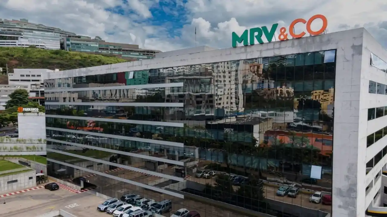 Fotografia aérea em plano geral e ângulo diagonal da fachada frontal do edifício-sede da MRV&CO. O prédio possui uma arquitetura corporativa moderna com estrutura curva revestida por amplas janelas de vidro escuro espelhado, que refletem as nuvens do céu e o cenário urbano ao redor. No topo da cobertura branca, destaca-se o letreiro institucional com a marca "MRV&CO" em letras garrafais nas cores verde e laranja. Em primeiro plano, há um estacionamento descoberto com fileiras de carros e, ao fundo, encostas com vegetação e outros edifícios urbanos sob um céu parcialmente nublado.