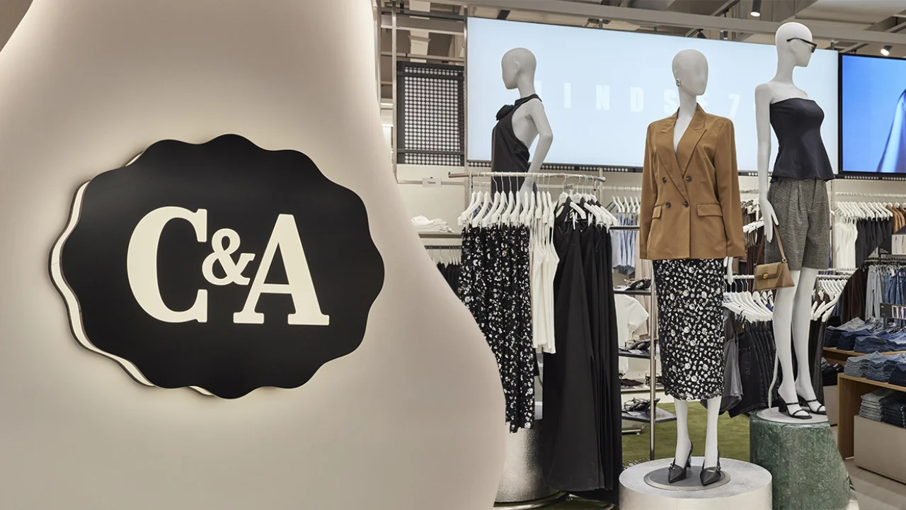 Interior de uma loja da C&A bem iluminada, destacando em primeiro plano um grande elemento decorativo curvo em tom neutro com a emblemática placa preta e branca da C&A. Ao fundo, manequins com blazer bege, saia estampada e regata escura, ladeados por araras de roupas femininas e telas digitais da loja.