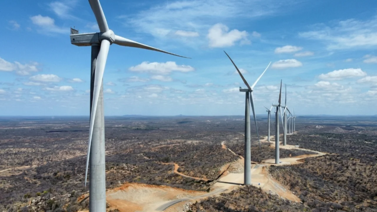 Vista aérea em perspectiva de um parque eólico da Auren Energia em terreno semiárido, mostrando uma sequência de altos aerogeradores brancos com três pás posicionados ao longo de uma estrada de terra, sob céu azul com nuvens dispersas.