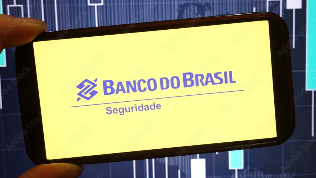 Mão segurando na horizontal um smartphone com tela amarela brilhante exibindo o logotipo azul do "BANCO DO BRASIL Seguridade". Ao fundo, desfocado, observa-se o painel escuro de um monitor com gráficos financeiros de velas e barras da Bolsa de Valores.