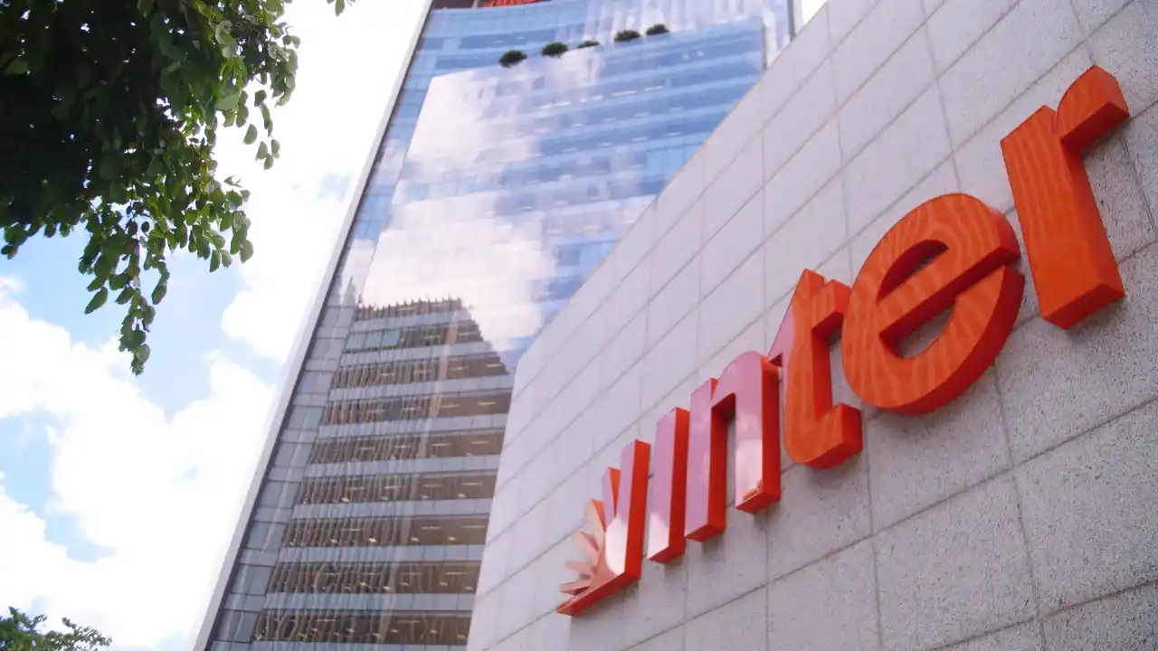 Fachada corporativa moderna do Banco Inter, mostrando em ângulo inferior o grande logotipo em alto-relevo na cor laranja fixado em parede de pedras cinza. Ao fundo, um arranha-céu com fachada de vidro espelhado reflete o céu azul com nuvens, ladeado por folhagens verdes no canto superior esquerdo.
