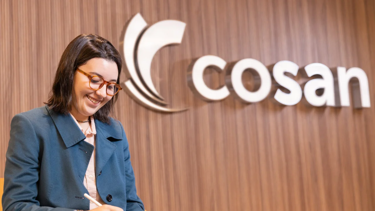 Profissional em escritório corporativo diante da logomarca oficial da Cosan em painel de madeira.
