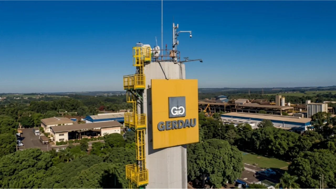 Vista aérea de uma unidade industrial da Gerdau, com destaque para uma grande torre de concreto em primeiro plano exibindo uma placa amarela com o logotipo prateado e o nome "GERDAU". Ao fundo, sob céu azul limpo, observam-se galpões de produção siderúrgica, maquinários, estruturas industriais e áreas verdes.