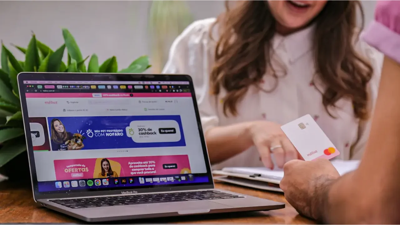 Mão em primeiro plano segurando o cartão de crédito branco do Méliuz com detalhes rosa e amarelo, em frente a um notebook aberto mostrando o site da plataforma com banners de cashback, com mulher ao fundo sorrindo em ambiente bem iluminado.