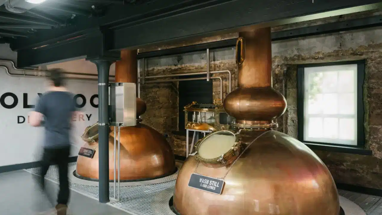 Interior de uma destilaria com dois grandes alambiques de cobre em primeiro plano. Há tubulações metálicas, estrutura industrial escura, paredes de pedra e uma janela ao fundo. Uma pessoa desfocada caminha à esquerda da imagem