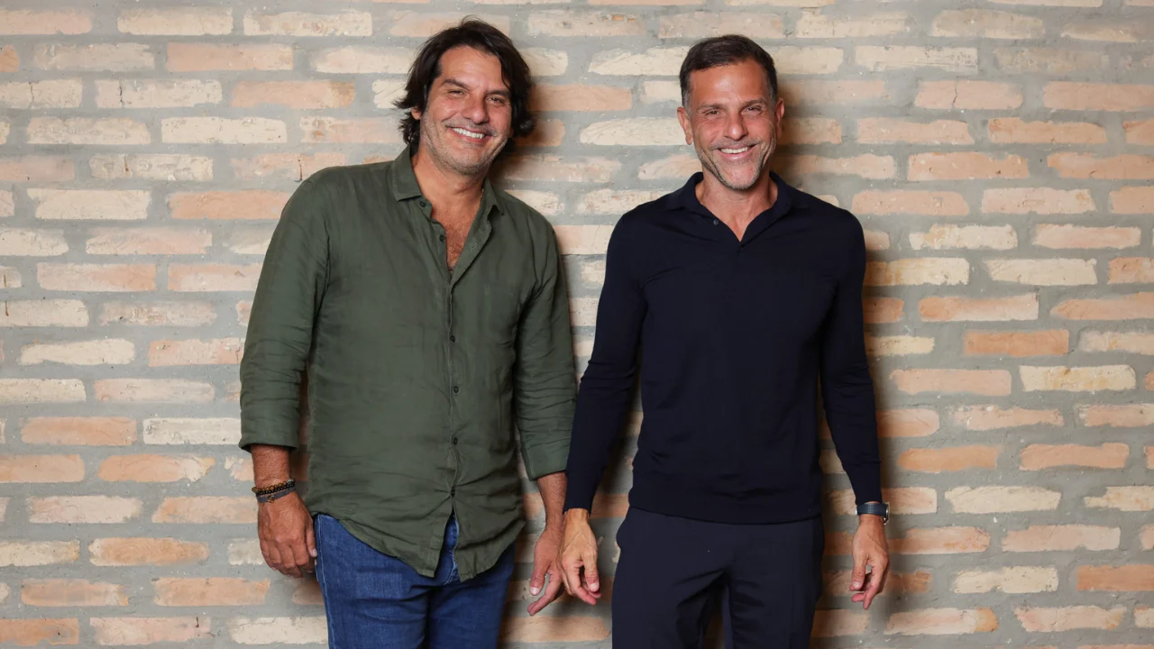 Roberto Jatahy e Alexandre Birman sorrindo em frente a uma parede de tijolos aparentes.