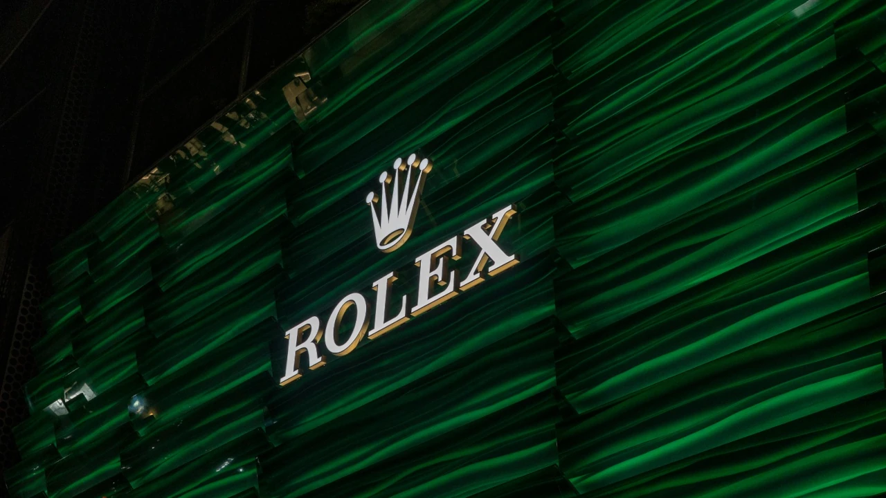 Logotipo da Rolex iluminado em destaque sobre uma fachada moderna com painéis texturizados em tons de verde escuro.