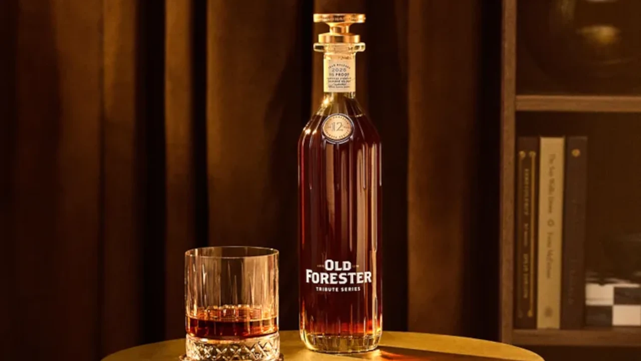 Garrafa de uísque Old Forester posicionada sobre uma mesa redonda ao lado de um copo com a bebida servida. Ao fundo, cortinas em tons amadeirados e uma estante com livros criam uma atmosfera sofisticada e intimista