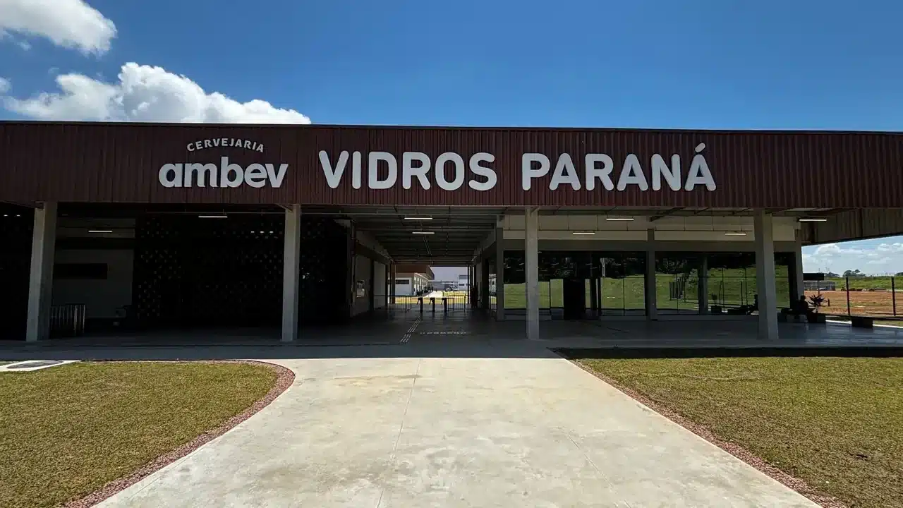Fachada da unidade industrial Cervejaria Ambev Vidros Paraná sob céu ensolarado, com caminho de concreto na entrada e gramados ao redor.