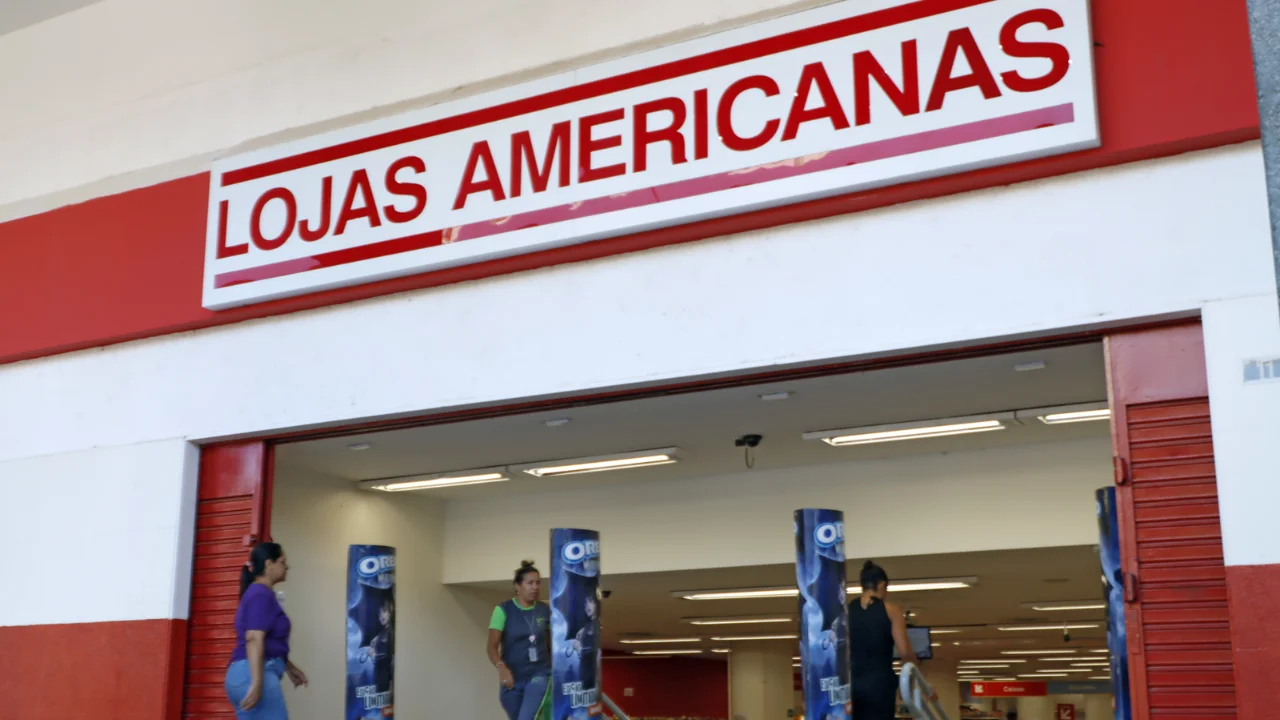 Fachada de entrada de uma unidade física das Lojas Americanas com o letreiro nas cores vermelha e branca em destaque, enquanto clientes caminham no local.