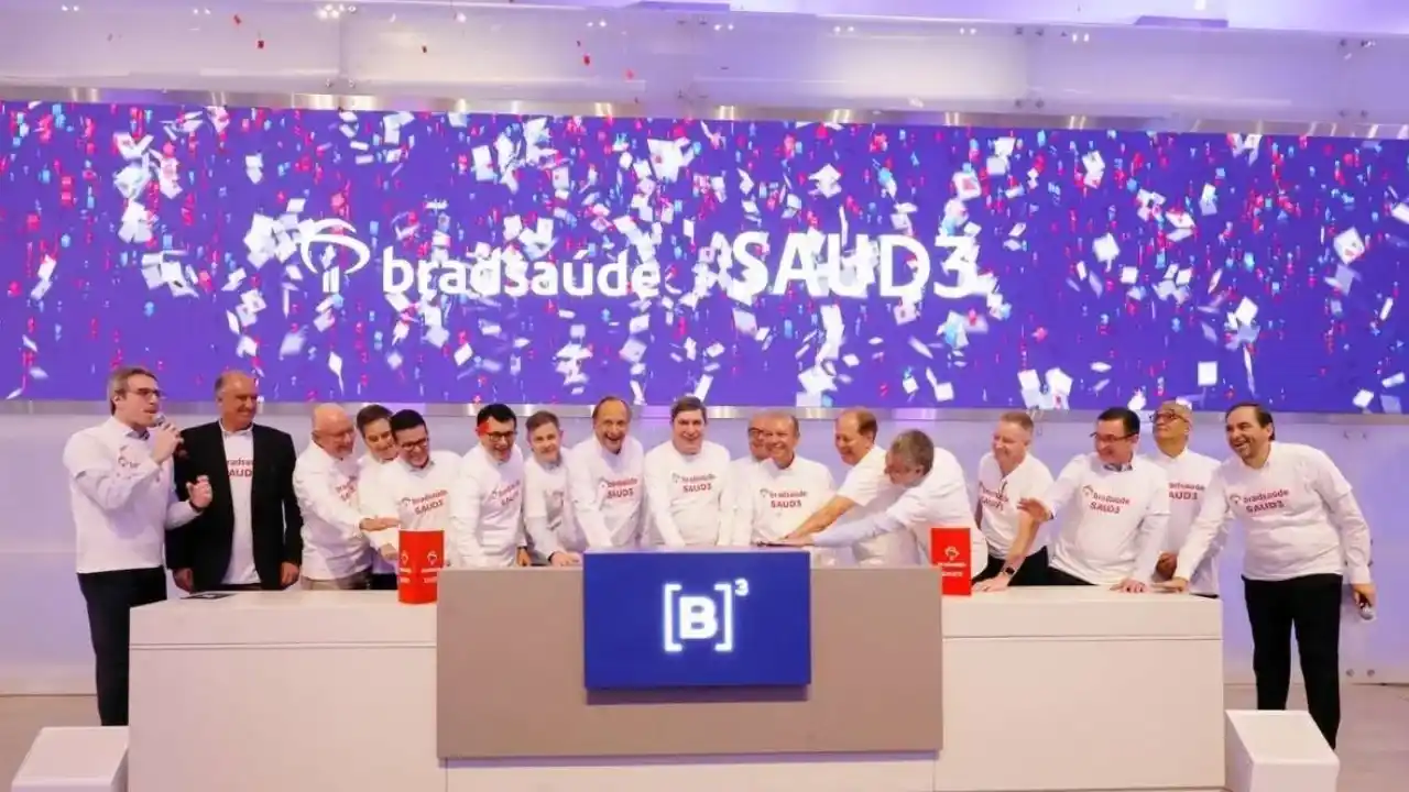 Executivos vestindo camisetas brancas com o logotipo da Bradsaúde reunidos no palco da B3 durante a cerimônia de estreia da empresa na bolsa, sob chuva de confetes e com um grande painel azul ao fundo exibindo a marca e o código SAUD3.