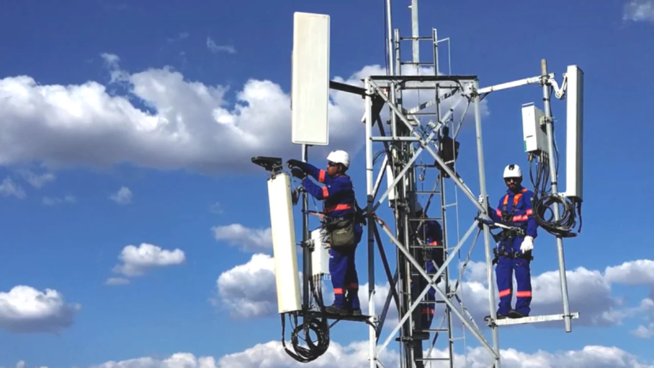 TÉCNICOS DA BRISANET TRABALHANDO EM UMA TORRE DE TELECOMUNICAÇÕES E ANTENAS DE TELEFONIA MÓVEL CONTRA UM CÉU AZUL COM NUVENS, ILUSTRANDO O CRESCIMENTO DOS ACESSOS MÓVEIS DA COMPANHIA.