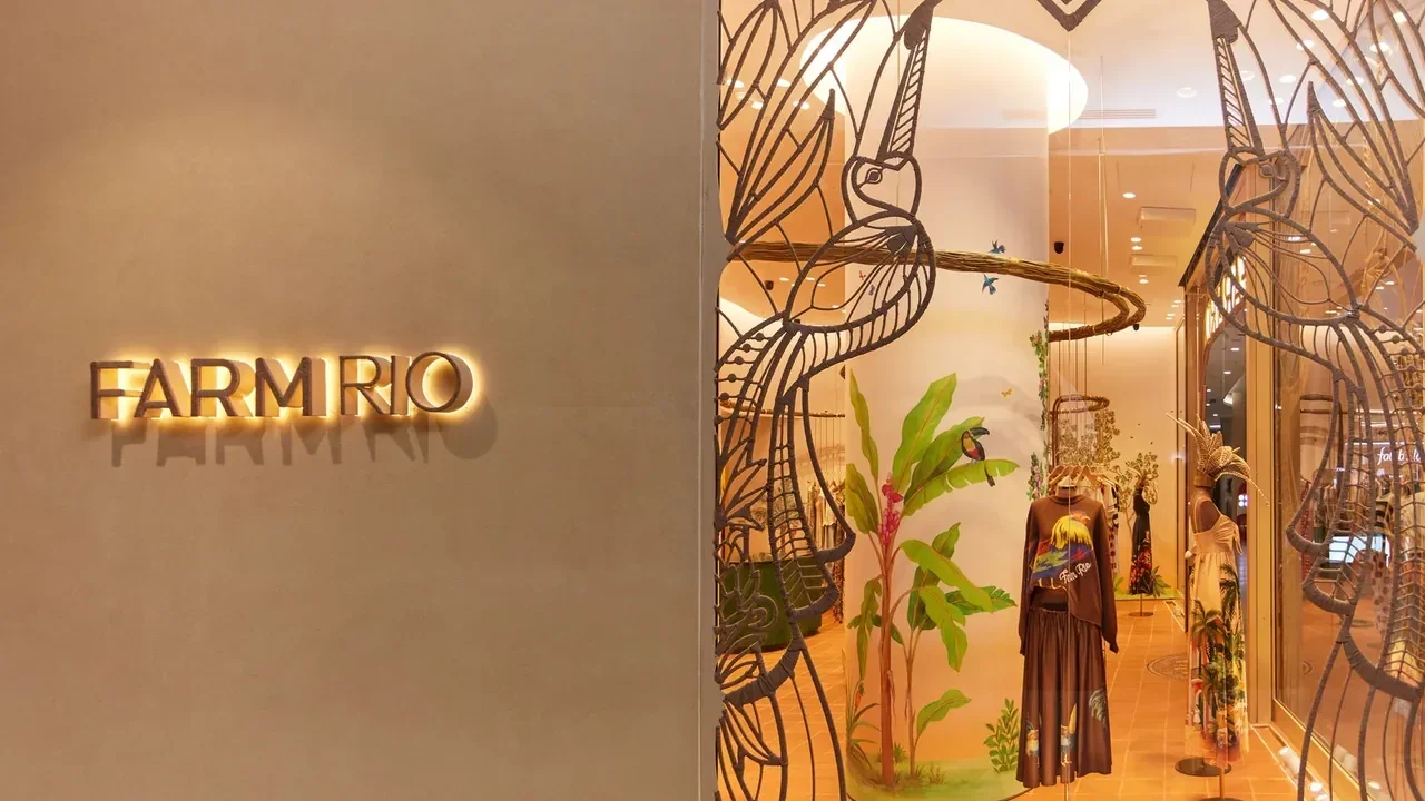 Fachada de loja com o nome Farm Rio iluminado em dourado em uma parede bege, ao lado de vitrine decorada com estruturas vazadas, estampas tropicais e manequins vestindo a coleção da marca