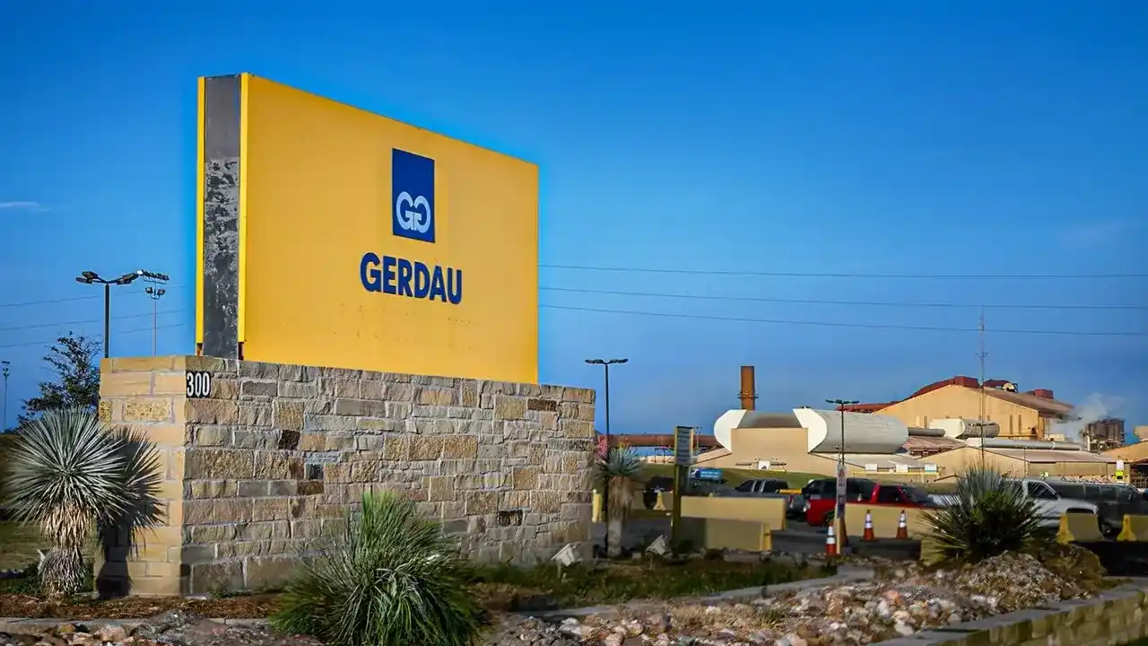 Placa amarela de entrada com a logo azul e o nome da Gerdau sobre base de pedra, na entrada de um complexo industrial siderúrgico sob céu azul.
