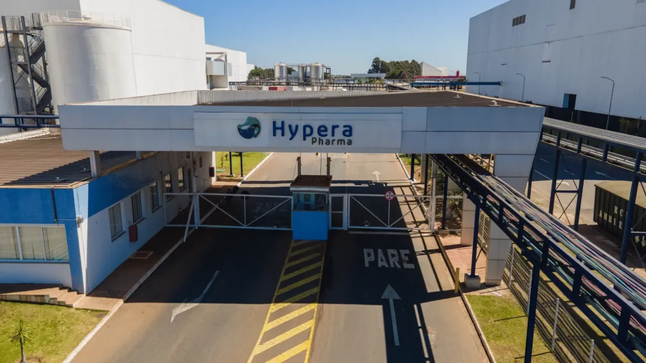 Portaria e fachada do complexo industrial da Hypera Pharma com guarita, sinalização de trânsito no asfalto e grandes edifícios fabris ao fundo sob céu azul.