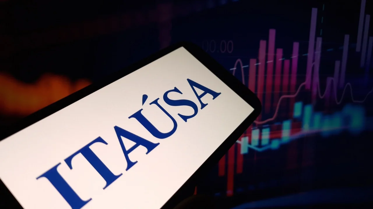Mão segurando um smartphone que exibe o logotipo da Itaúsa em destaque, com tela de gráfico de cotações de fundo em tons escuros e coloridos, ilustrando a análise do desconto de holding.
