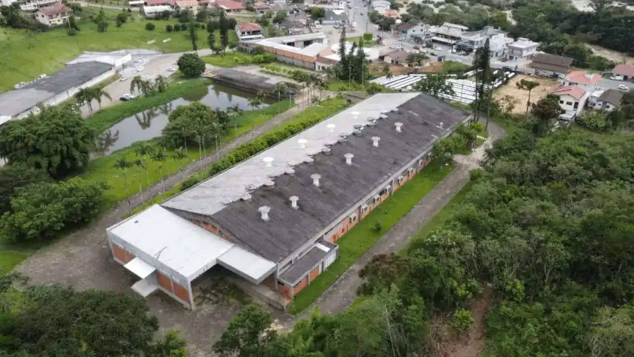 Vista aérea de galpão industrial retangular com telhado escuro e saídas de ar, cercado por vegetação densa, áreas verdes e vias urbanas ao fundo em Ibirama, Santa Catarina.