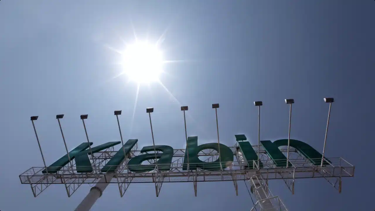 Letreiro estruturado em letras verdes com a palavra Klabin visto de baixo para cima contra o céu azul com o sol reluzindo ao fundo.