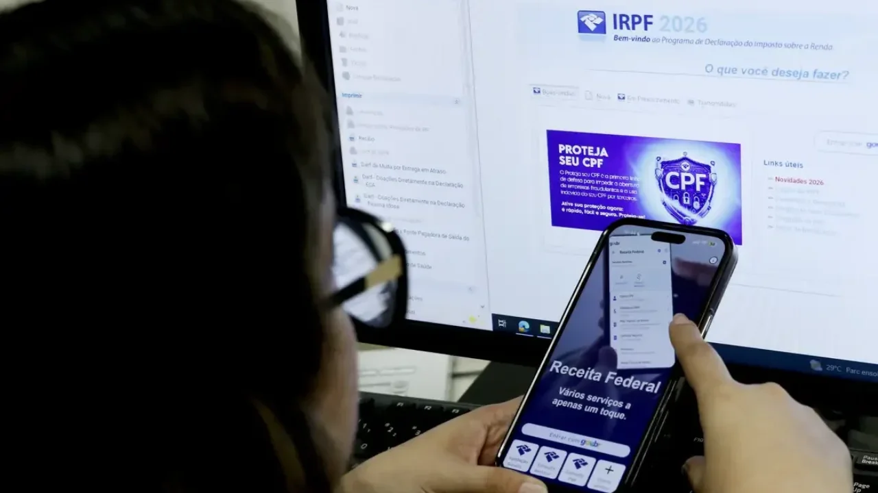 Contribuinte usa o celular e computador com a tela do IRPF 2026 para consultar extrato da malha fina e e-CAC da Receita Federal.