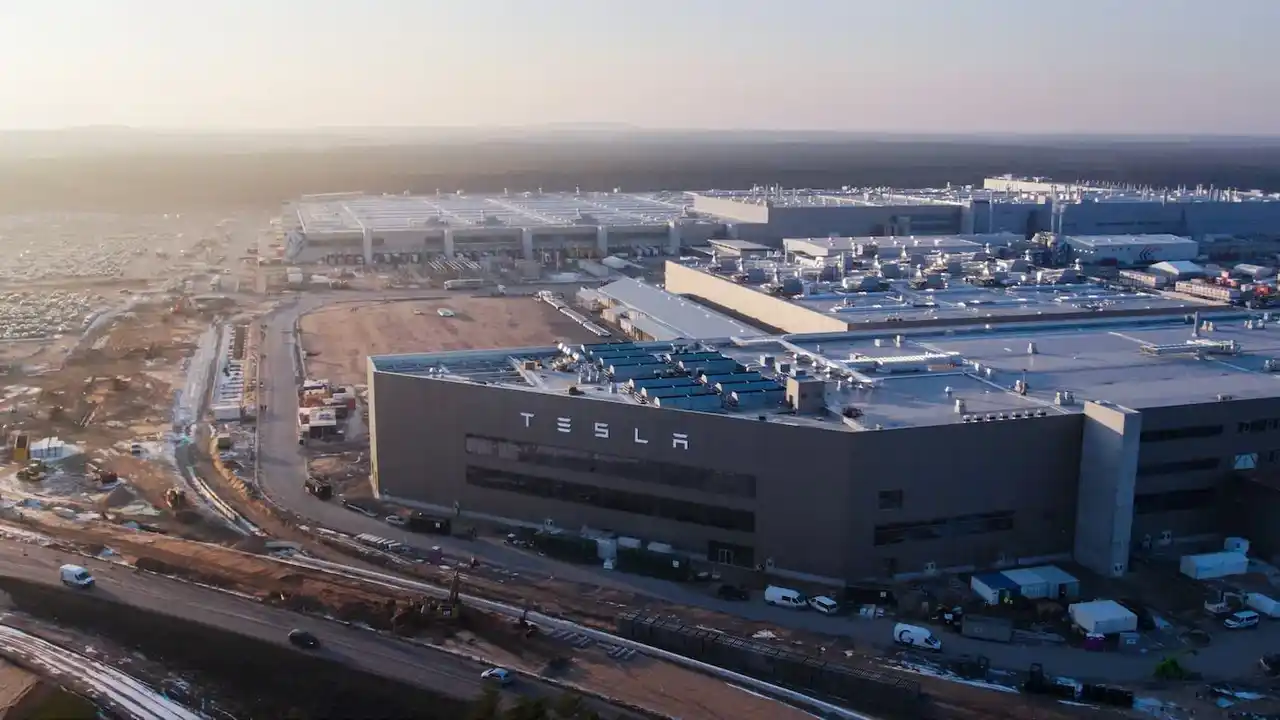 Vista aérea panorâmica do gigantesco complexo industrial da Gigafactory da Tesla na Alemanha, destacando a fachada com a palavra Tesla em destaque, o pátio de estacionamento e as vias de acesso no entorno ao nascer do sol.