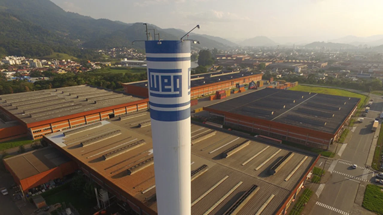 Vista aérea do parque fabril da WEG com grande chaminé e reservatório industrial vertical exibindo o logotipo da WEG em azul e branco, cercado por galpões de produção e relevo montanhoso ao fundo.