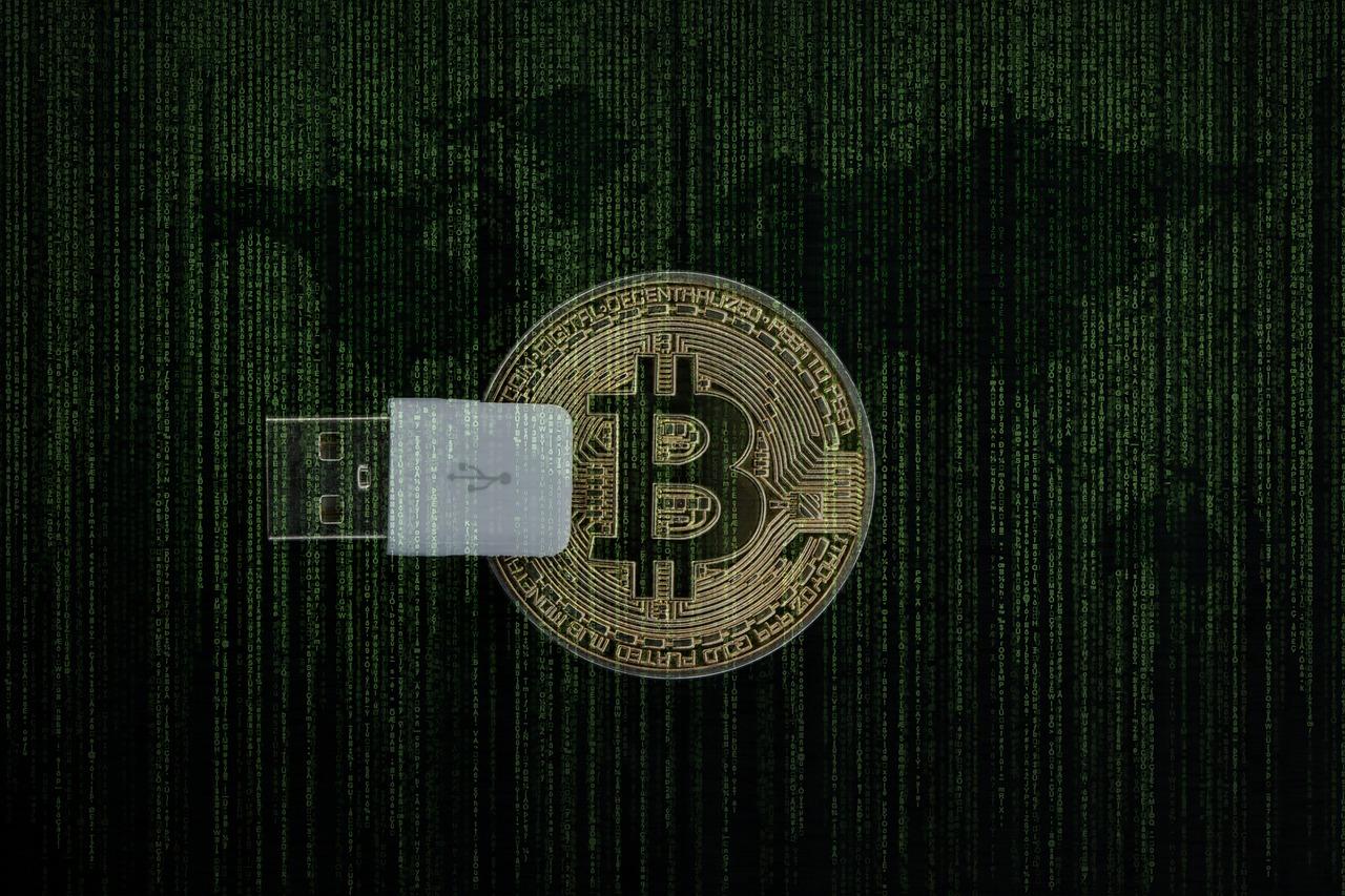A imagem mostra um Bitcoin com um pendrive de blockcahin e um quadro de programação de fundo.