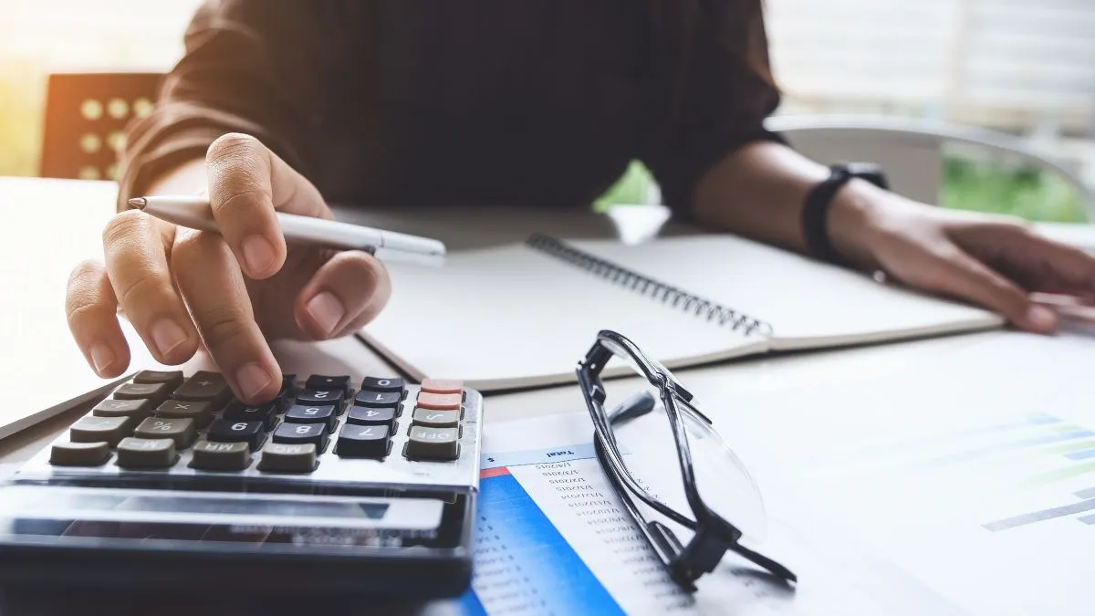 Pessoa usando uma calculadora em uma mesa com cadernos e óculos, ilustrando a análise de investimentos ou finanças